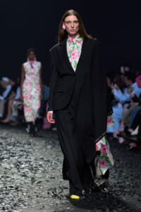 Model walks the runway in an oversized black suit over a pink-and-green floral blouse, with a pink heart-shaped earring and a yellow shoe tip visible at the front.
