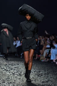 Model on a dark runway wearing an all-black outfit with a large sculptural hat and a Moschino belt, hands in pockets as confetti falls on the floor.