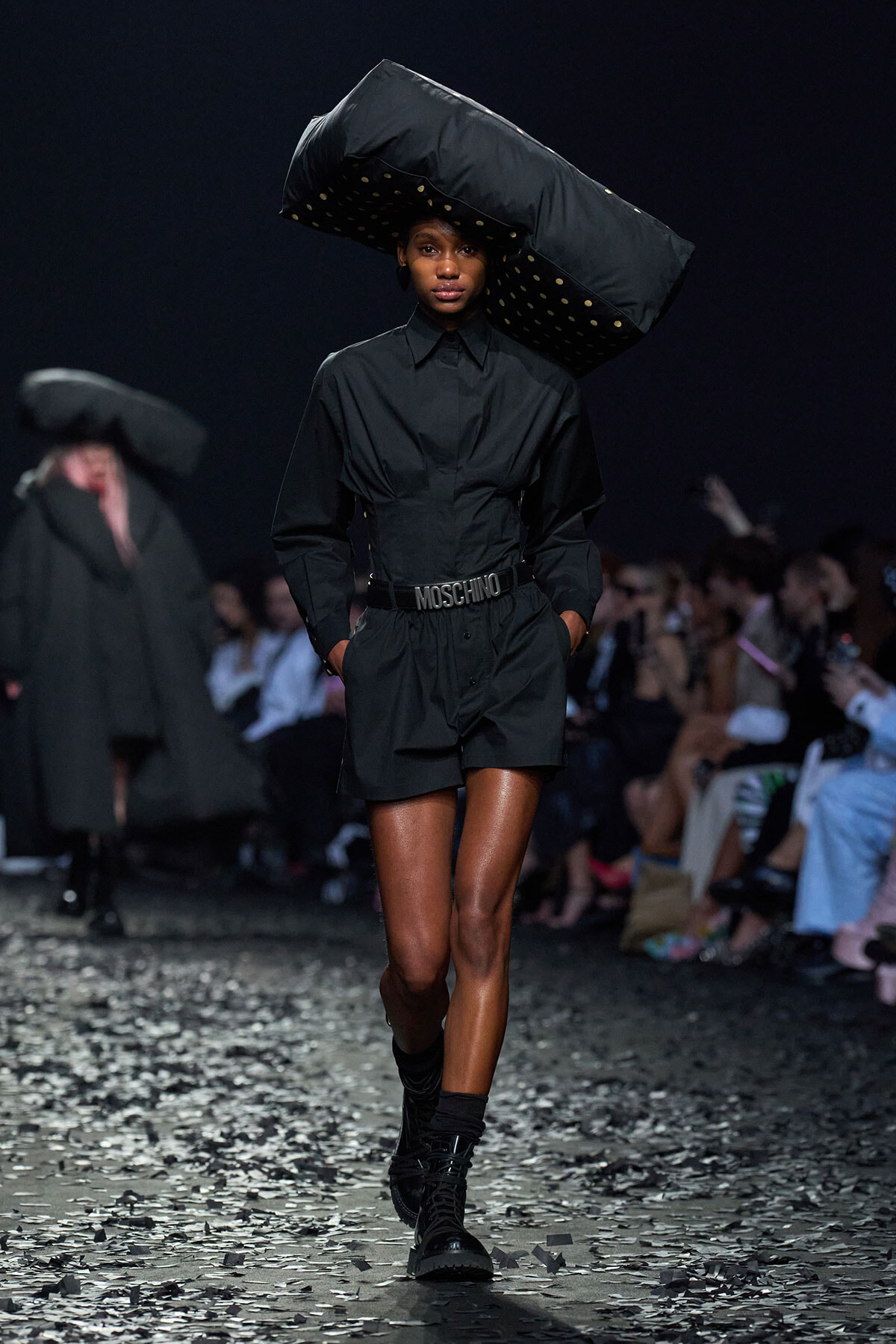 Model on a dark runway wearing an all-black outfit with a large sculptural hat and a Moschino belt, hands in pockets as confetti falls on the floor.