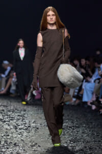Model on a fashion runway wearing a brown cold-shoulder dress-like top with matching pants, gloves, and a fluffy gray fur bag.