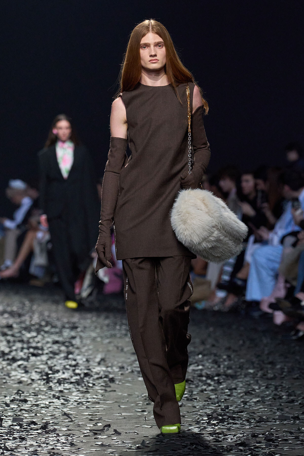 Model on a fashion runway wearing a brown cold-shoulder dress-like top with matching pants, gloves, and a fluffy gray fur bag.