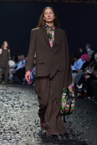 Model walks a fashion runway wearing a oversized brown suit with a bright floral blouse; she carries a matching floral bag as confetti falls on the catwalk.