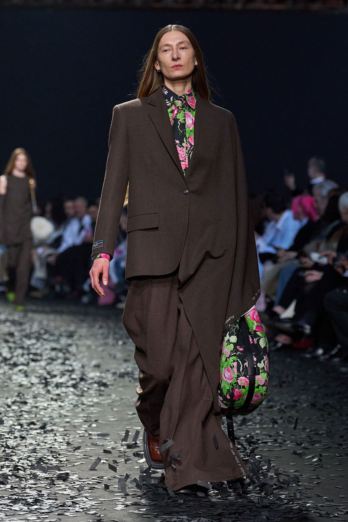 Model walks a fashion runway wearing a oversized brown suit with a bright floral blouse; she carries a matching floral bag as confetti falls on the catwalk.
