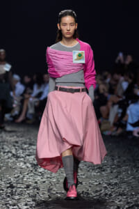 Model walking a runway in a pink and gray outfit with a pink scarf and glossy pink shoes, confetti on the floor.