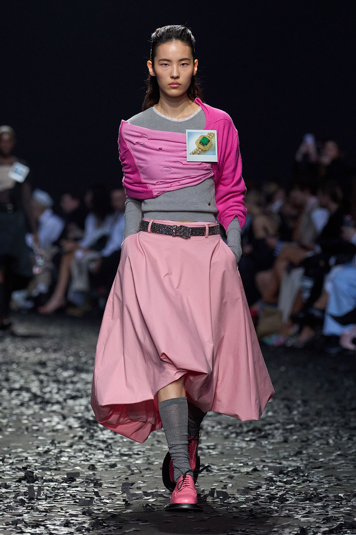Model walking a runway in a pink and gray outfit with a pink scarf and glossy pink shoes, confetti on the floor.