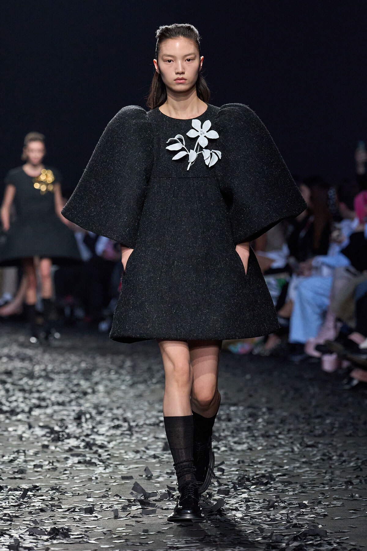 Runway model wearing a black textured mini dress with oversized cape sleeves and a white floral brooch, walking on a confetti-strewn runway.