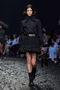 Model walking a runway in a black blouse with oversized bow and black polka-dot-hem skirt, wearing a Moschino belt and carrying a black stuffed dog purse.