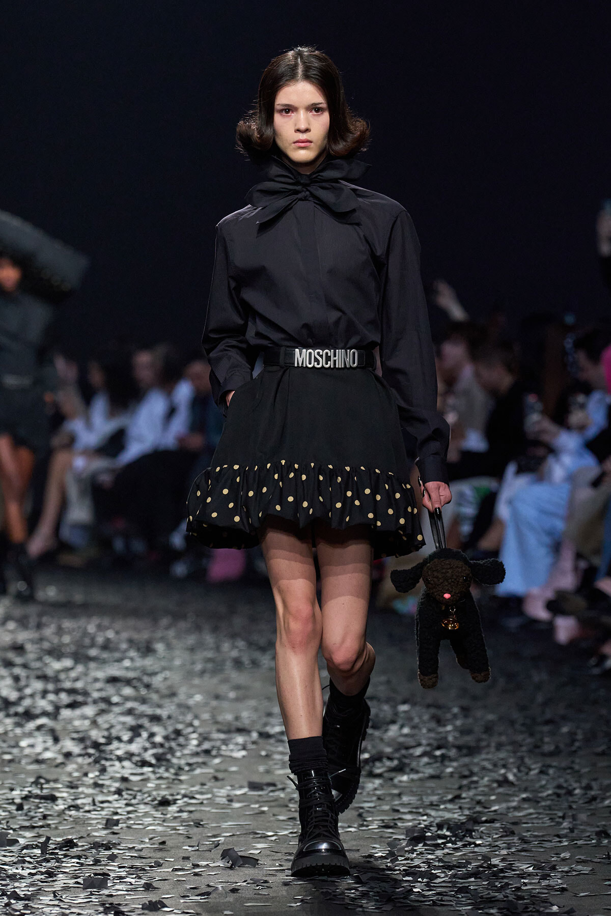 Model walking a runway in a black blouse with oversized bow and black polka-dot-hem skirt, wearing a Moschino belt and carrying a black stuffed dog purse.