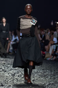 Runway model in a brown and beige wrapped top with a voluminous black skirt, brown boots, and a small card pinned to the chest. Audience watches from the background.