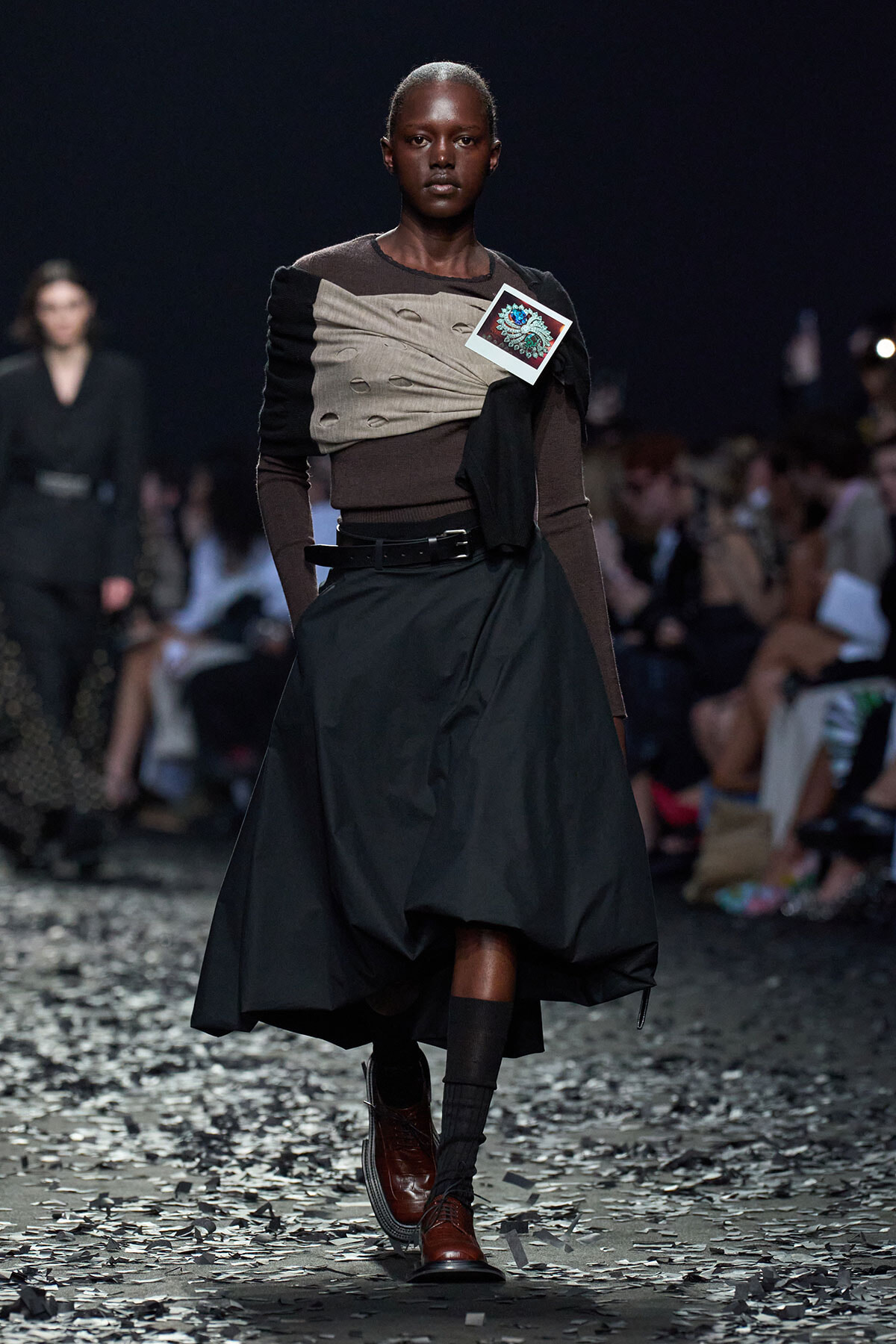 Runway model in a brown and beige wrapped top with a voluminous black skirt, brown boots, and a small card pinned to the chest. Audience watches from the background.
