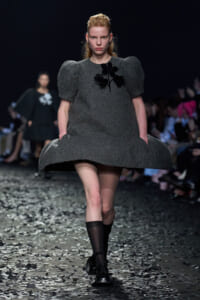 Model on a runway wearing a dark gray textured dress with oversized puff sleeves and a black flower applique on the chest.