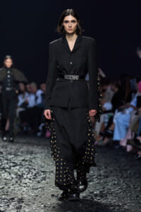 Female model walking a runway in a tailored black suit with a studded Moschino belt and polka-dotted ruffled panels, confetti on the floor, dark backdrop, audience in the background.