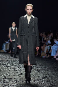Model on a fashion show runway wearing a dark gray, tailored long coat with white contrast stitching and a cream turtleneck underneath.