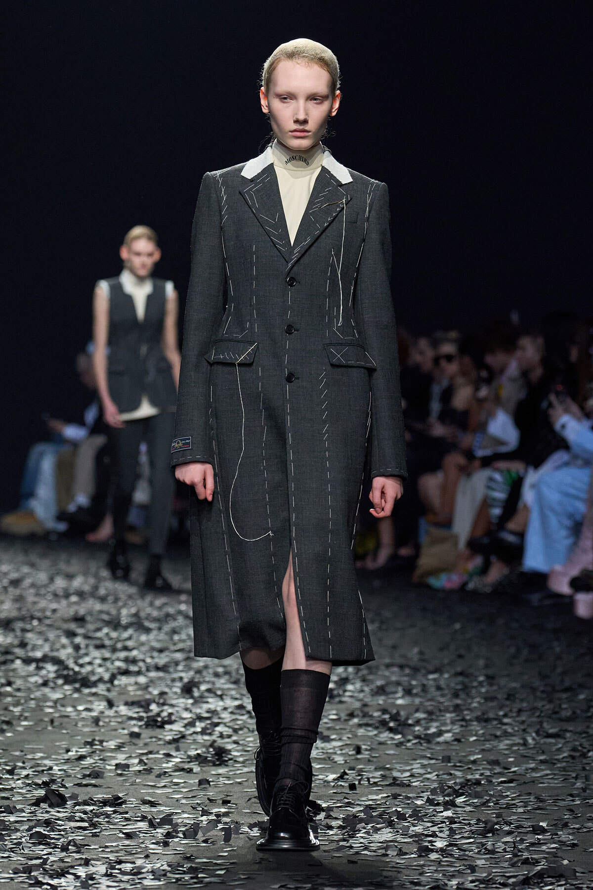 Model on a fashion show runway wearing a dark gray, tailored long coat with white contrast stitching and a cream turtleneck underneath.