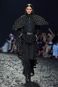 Model walks a black Moschino ensemble with oversized polka-dot cape, woven braids, and chunky belt on a confetti-strewn runway.