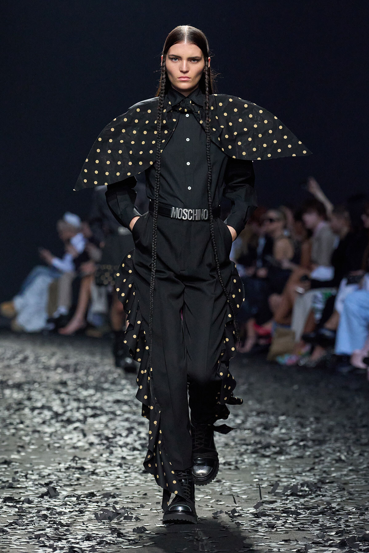 Model walks a black Moschino ensemble with oversized polka-dot cape, woven braids, and chunky belt on a confetti-strewn runway.
