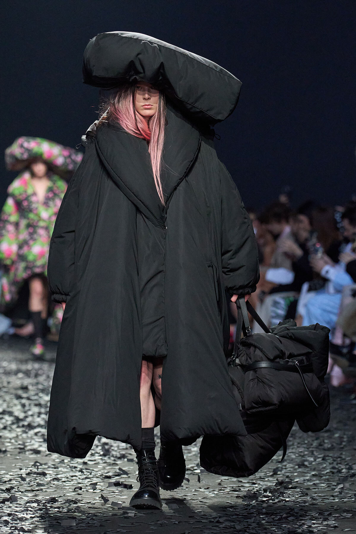 Model walks a runway in an oversized black puffer coat and a dramatic sculptural hat, carrying a large padded bag, with confetti on the floor.