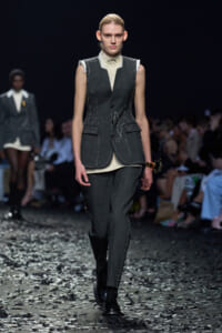Fashion model walks a runway in a dark gray tailored suit with a sleeveless vest, white high-collar top, and polished black boots, confetti on the floor.