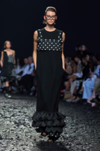 Model walking a fashion runway in a black gown with a polka-dotted overlay and tiered ruffled hem, wearing bold black glasses.