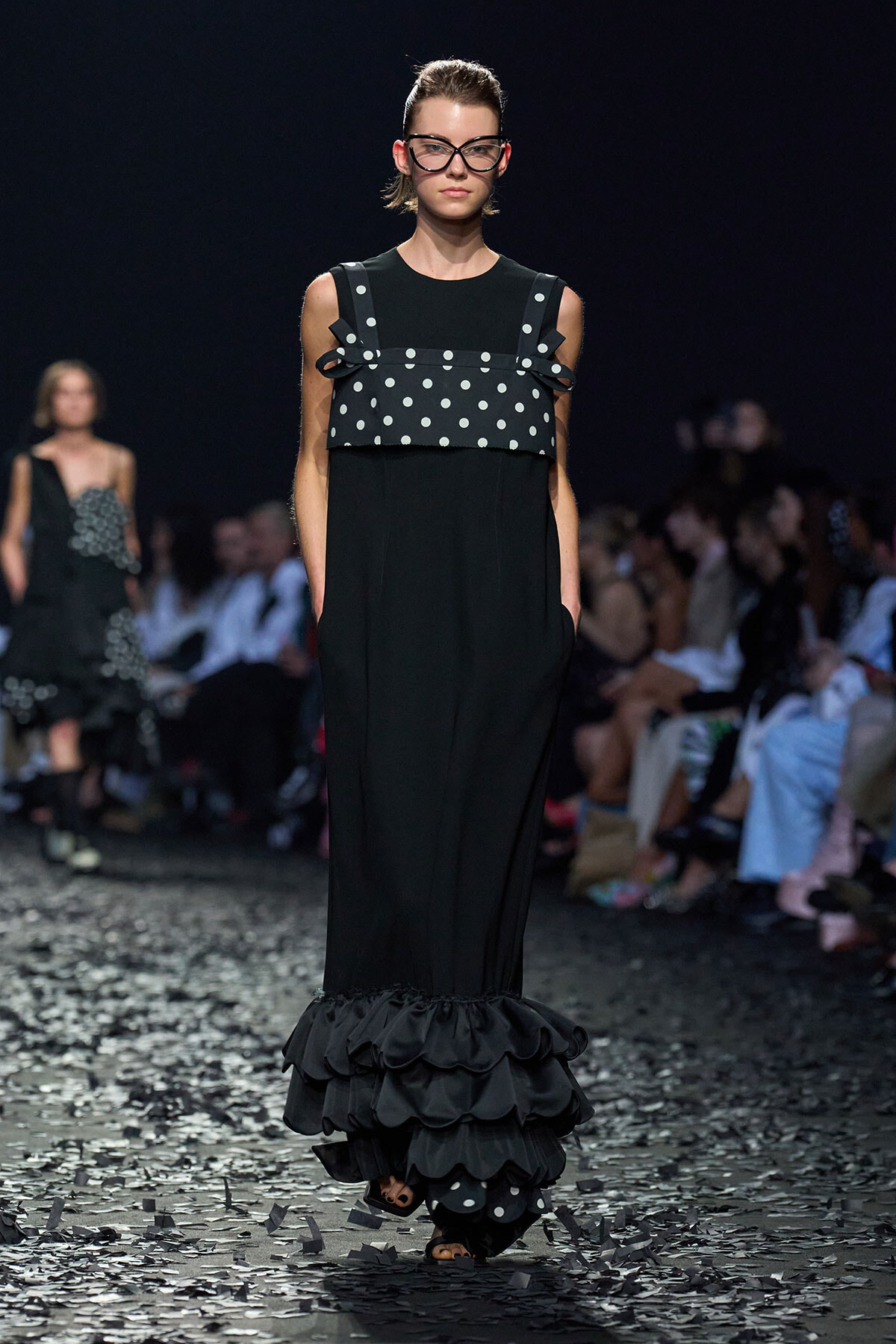 Model walking a fashion runway in a black gown with a polka-dotted overlay and tiered ruffled hem, wearing bold black glasses.