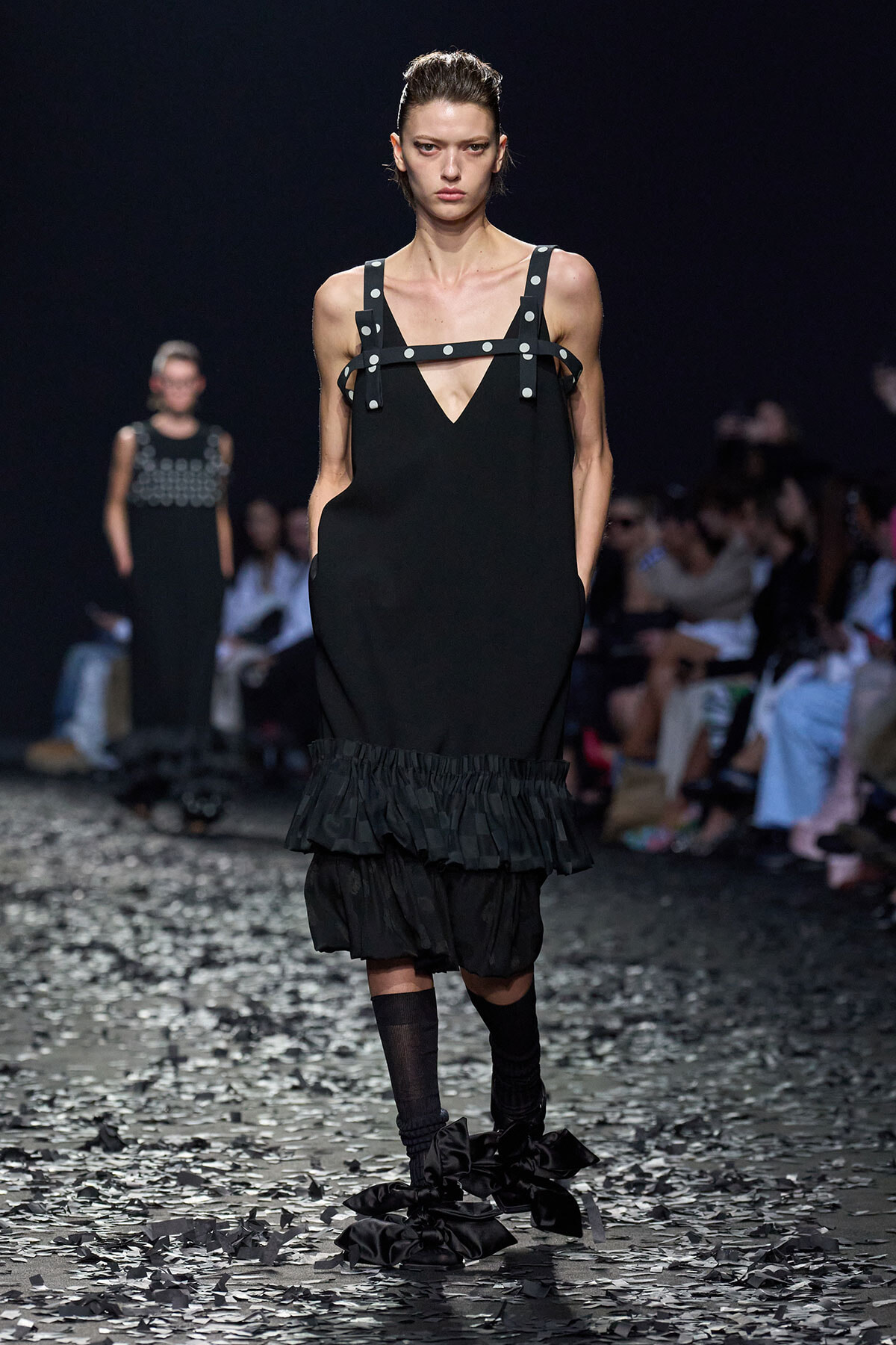 Model on a runway wearing a black V-neck dress with studded harness straps and a ruffled hem; audience blurred in background.