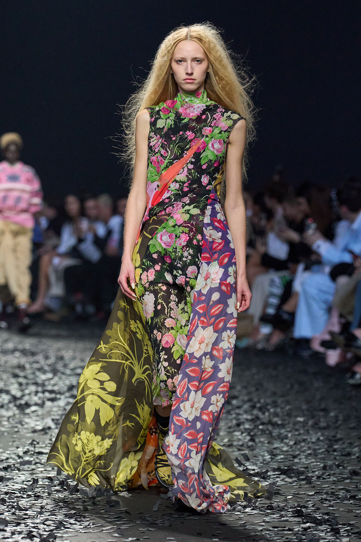 Model wearing a vibrant, multi-panel floral gown with a sheer train on a runway.