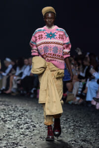 Model walking the runway in a pink patterned knit sweater, a tan oversized cargo wrap skirt, a tan beret, burgundy lace-up boots, and a small blue handbag; audience blurred in the background.