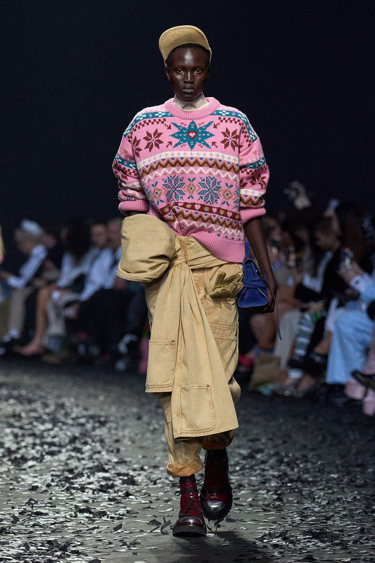 Model walking the runway in a pink patterned knit sweater, a tan oversized cargo wrap skirt, a tan beret, burgundy lace-up boots, and a small blue handbag; audience blurred in the background.