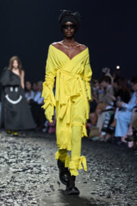 Runway model in a bright yellow wrap dress with multiple belts, wearing gloves, black boots, a head wrap, and dark sunglasses.