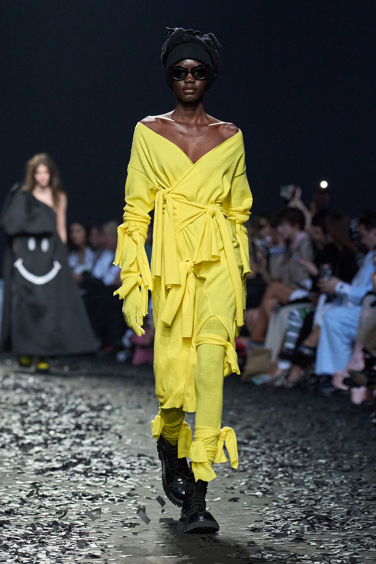 Runway model in a bright yellow wrap dress with multiple belts, wearing gloves, black boots, a head wrap, and dark sunglasses.