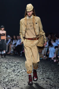 Runway model in a camel-toned outfit with cap, jacket, belt, and tan pants, holding a tan bag; shirt reads 'LUXURY IS RELATIVE'.