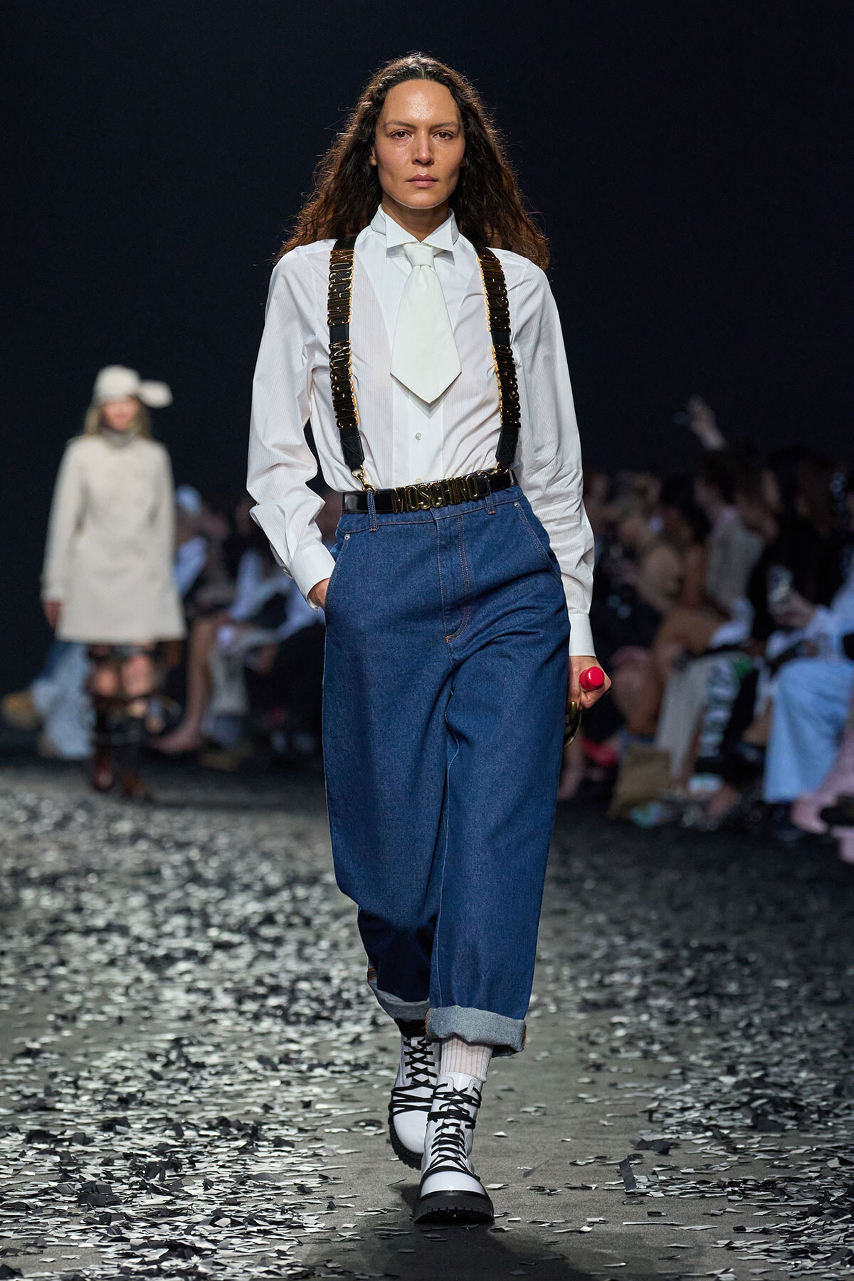 Model walks a runway in a white button-down shirt, pale tie, dark suspenders, and rolled-hem jeans with black-and-white lace-up boots.