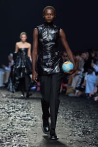 Model walks a fashion runway in a shiny black vest and black pants, holding a small globe, with audience and other models in the background.