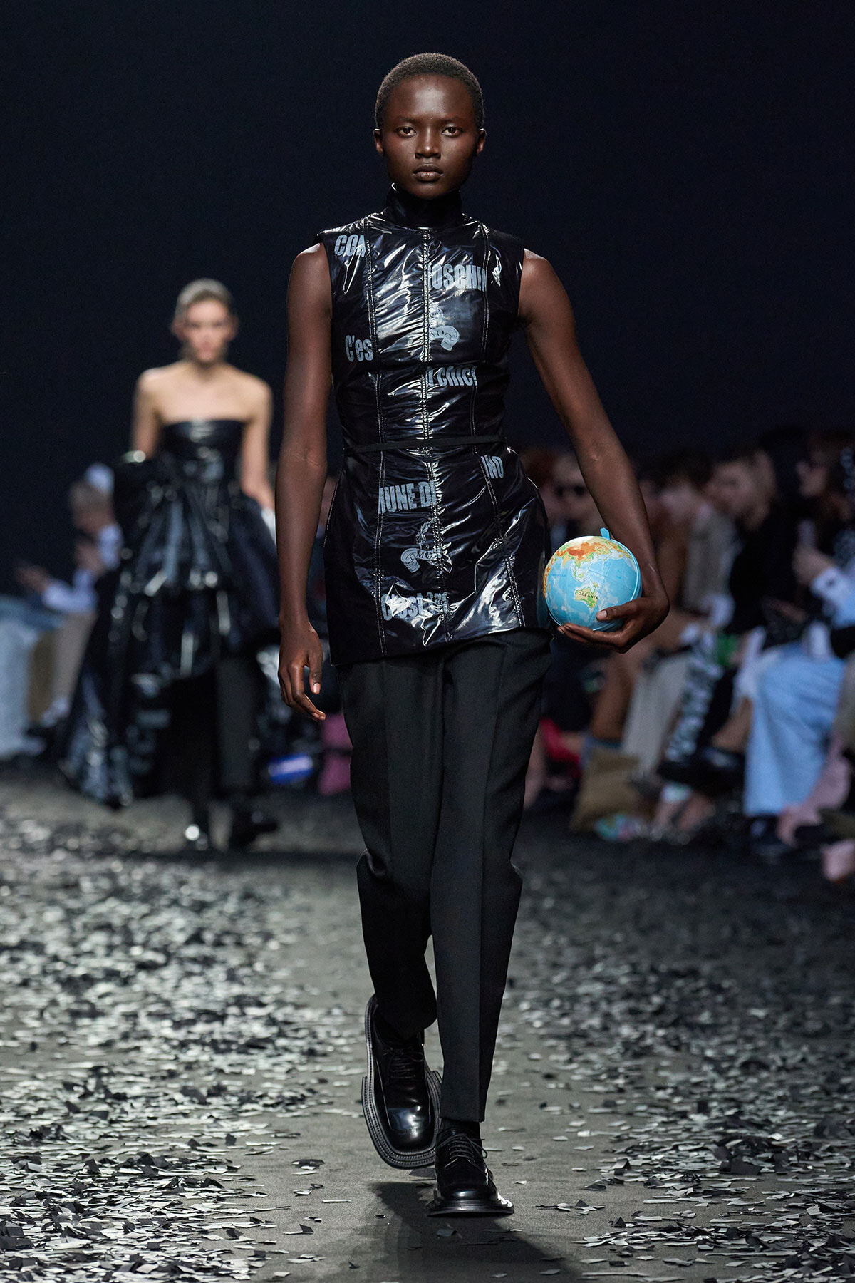 Model walks a fashion runway in a shiny black vest and black pants, holding a small globe, with audience and other models in the background.