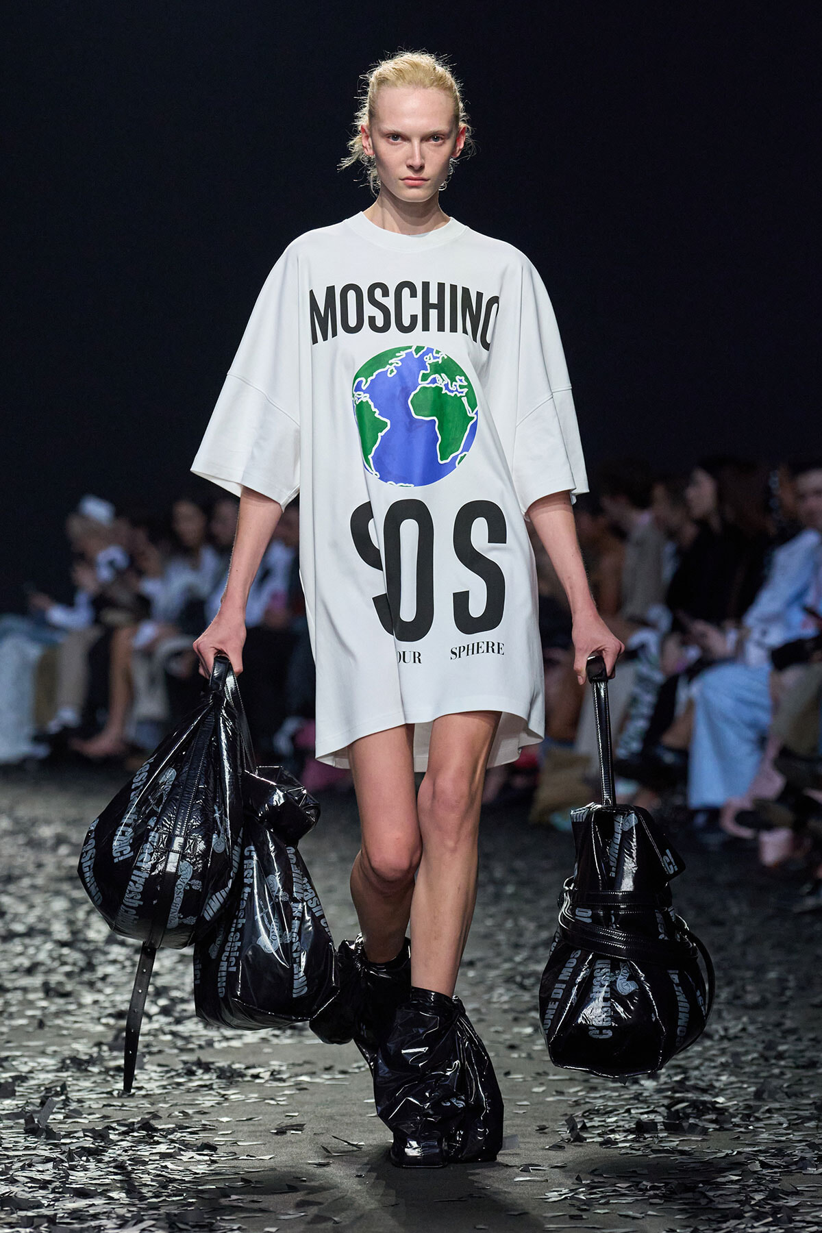 Runway model in a white oversized Moschino T-shirt with a globe graphic and bold MOSCHINO/SOS lettering, carrying black bags.