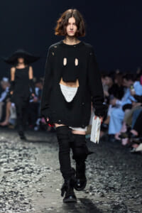Model walks a runway in an oversized black knit sweater with intentional rips and cutouts, showing white underlayer, paired with black thigh-high leg warmers and combat boots, holding a white folder or booklet backstage prop.