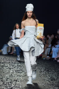 Model in an all-white, off-shoulder dress and chef hat walks a runway while holding a yellow-and-white Moschino cookie box.