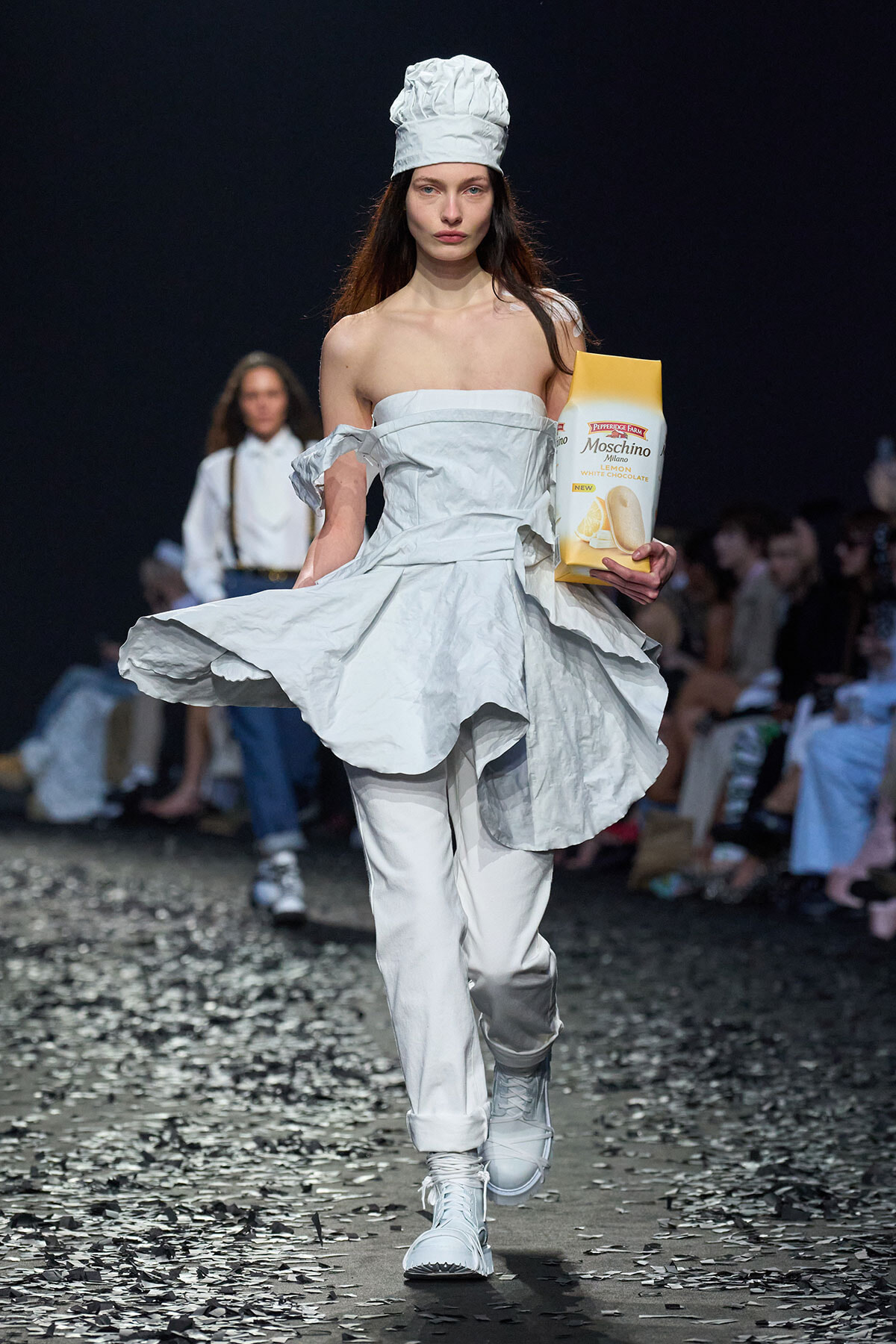 Model in an all-white, off-shoulder dress and chef hat walks a runway while holding a yellow-and-white Moschino cookie box.