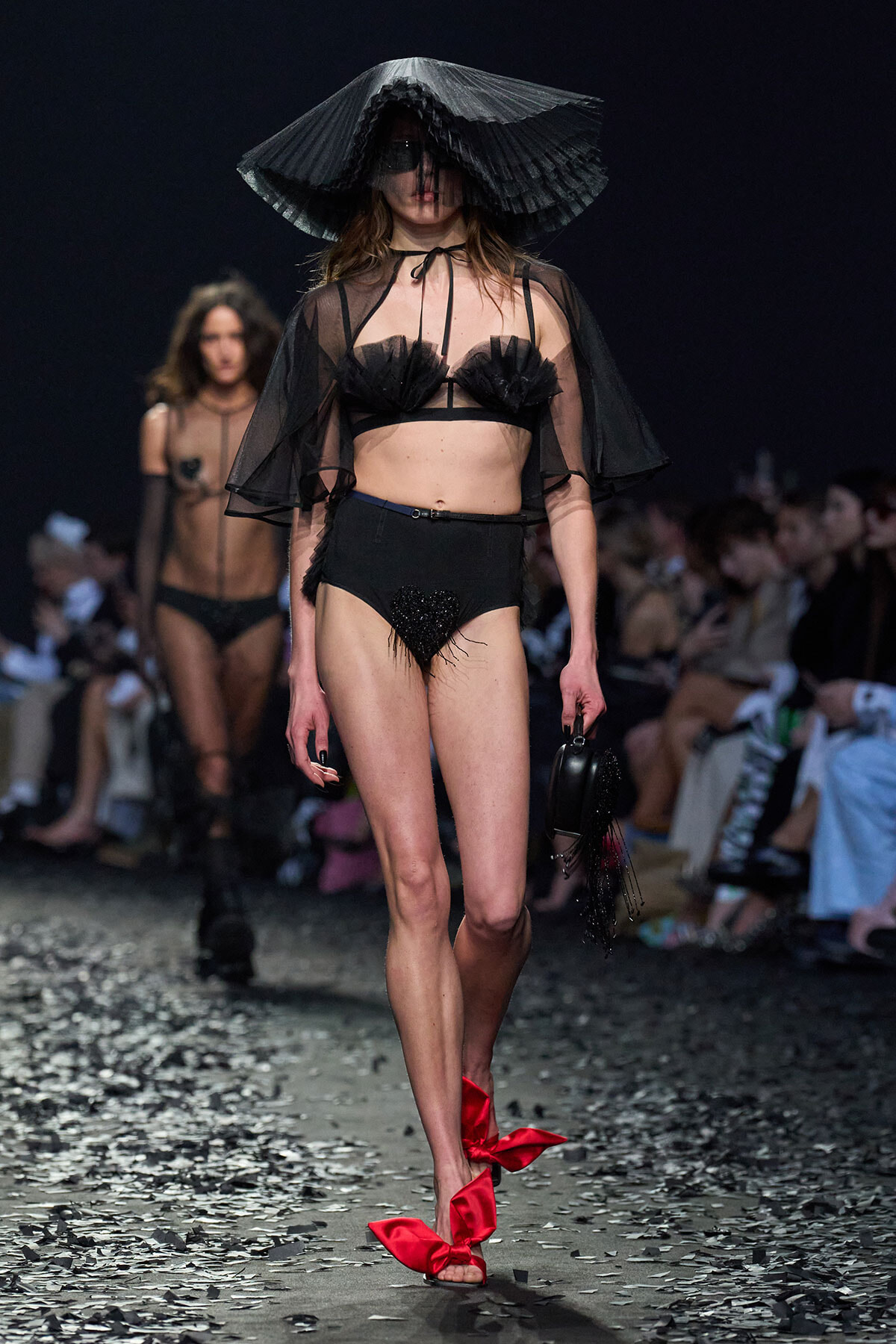 Runway model in black sheer lingerie with a large pleated hat, cape, and red ribbon sandals walking the catwalk with an audience in the background.