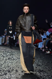 Model on a runway in a dark brown leather zip jacket, cap, and patchwork denim pants with an orange label; carrying a dark tote bag on the shoulder.