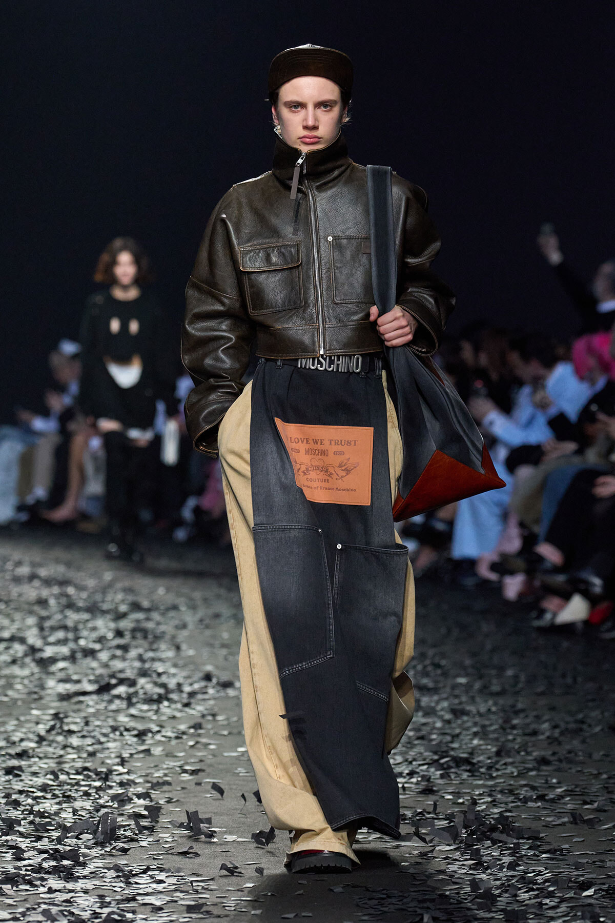 Model on a runway in a dark brown leather zip jacket, cap, and patchwork denim pants with an orange label; carrying a dark tote bag on the shoulder.
