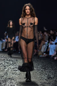 Model walking the runway in a sheer black mesh gown with heart pasties, long gloves, and black boots amid confetti.