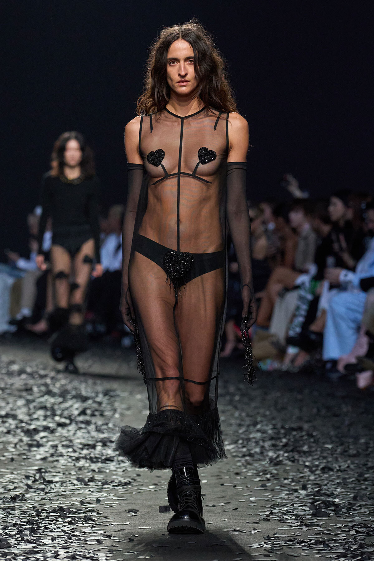 Model walking the runway in a sheer black mesh gown with heart pasties, long gloves, and black boots amid confetti.