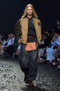 Model walks a runway wearing a tan varsity jacket with leather sleeves over a dark denim jumpsuit and red boots, audience in background.