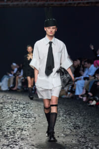 Runway model in an oversized white shirt, black tie, white shorts, black knee-high socks, and a tall black hat with an 'M', carrying a black bag.