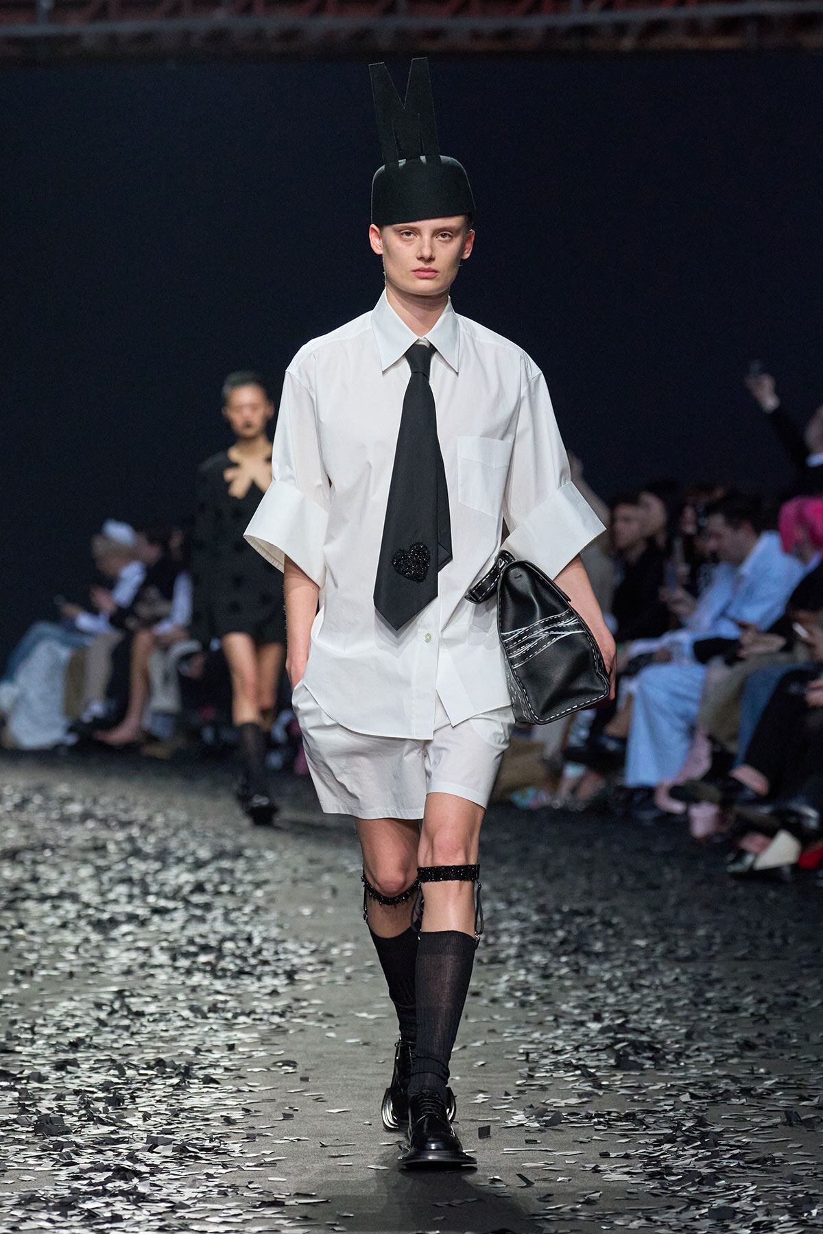 Runway model in an oversized white shirt, black tie, white shorts, black knee-high socks, and a tall black hat with an 'M', carrying a black bag.