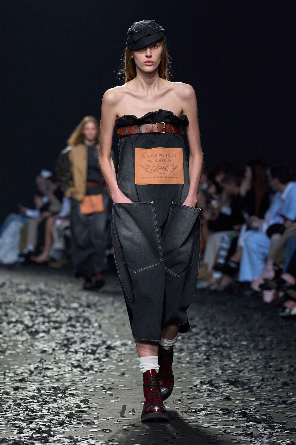 Runway model in a strapless dark denim jumpsuit with a brown belt, black hat, white socks, and red lace-up boots; large front patch visible on the jumpsuit.