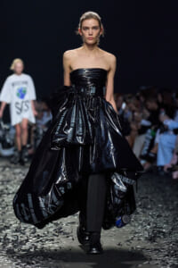 Model walking runway in a strapless, shiny black gown with oversized ruffles made from plastic-like material; dark audience and debris on the floor nearby.