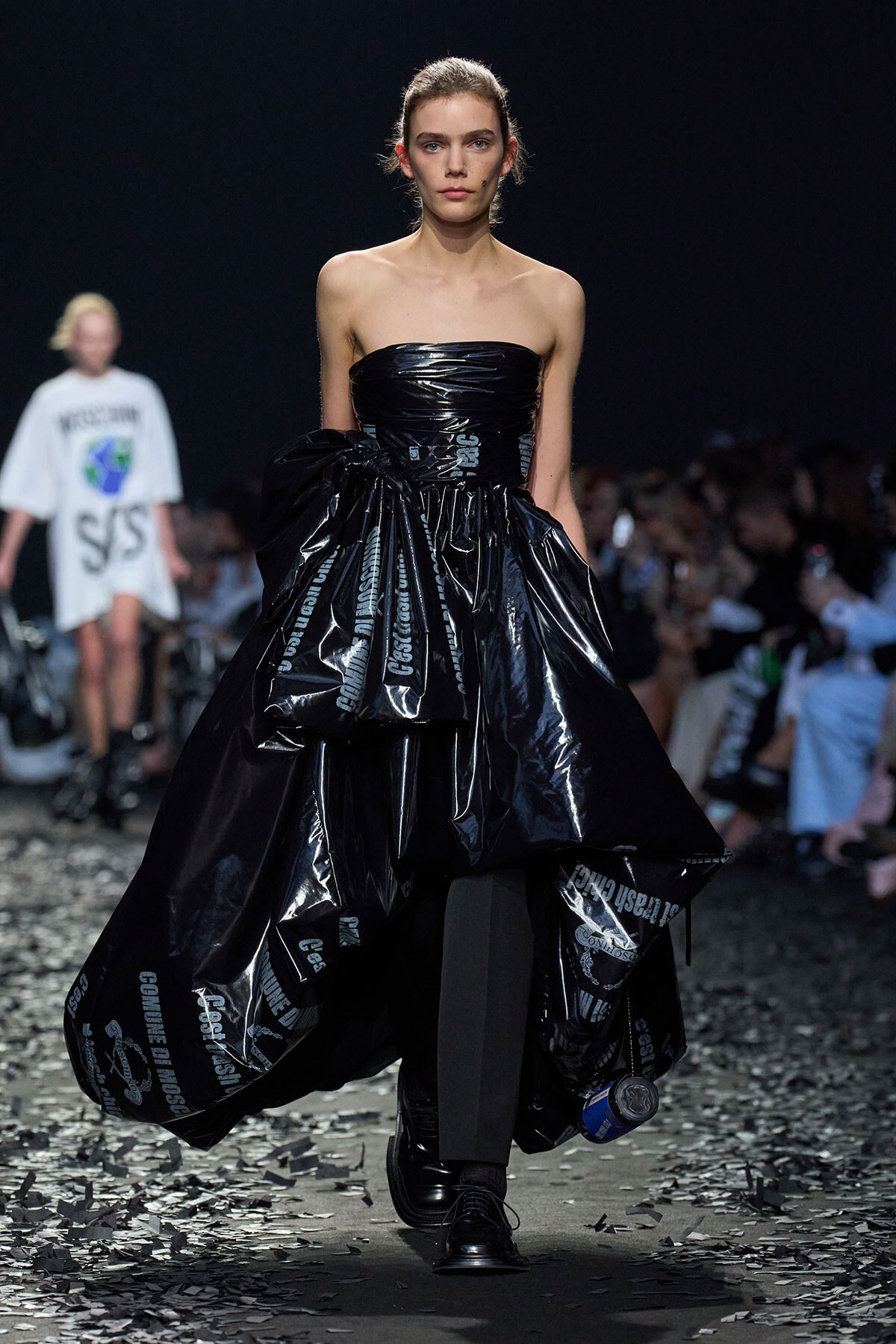 Model walking runway in a strapless, shiny black gown with oversized ruffles made from plastic-like material; dark audience and debris on the floor nearby.