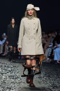 Model on a runway wearing a beige double-breasted trench coat with a sculptural hat and a leather leg harness over brown boots.